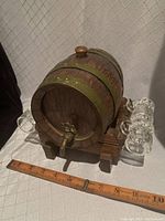 Full view of the oak mini keg with brass bands, brass spigot, and 4 glass shot mugs arranged around the oak stand, ruler for scale