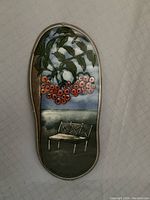 Front view of oval-shaped hand painted clay plaque featuring a scene with a bench and hanging branches with red berries under a blue sky with green ground.