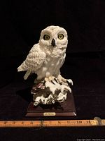 Front view of white owl sculpture showing detailed feathers, yellow eyes, perched on wood base with snow and E. Farina signature plaque.