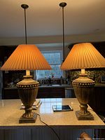 Pair of gold and black ceramic urn style table lamps placed side by side on a kitchen island, with pleated fabric shades that are illuminated and casting warm light.