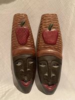 Front view of two carved wooden wall masks with painted apples on top and carved faces beneath.