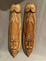 Front view of two elongated wooden masks with carved and painted facial features and white dot accents.