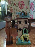 Photo showing the wooden cat figurine and folk art birdhouse side by side on a wooden floor with floral background.