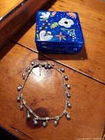 Photo showing the blue square ceramic trinket box with colorful floral paintings and the silver ankle bracelet with dangling bells on a wooden table.