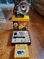 Kodak Brownie Hawkeye camera with large flash reflector mounted, original box beneath showing mother and child image, interior box includes flash bulbs and instruction sheet.