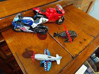 All four die cast miniatures displayed on a wooden surface: two motorcycles (red and blue), one black helicopter with red blades, one blue and white propeller airplane.