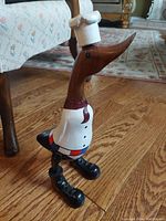 Side angle of the wooden hand carved and painted duck figurine in chef attire standing on hardwood floor beside furniture.