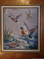 Frontal view of the vintage needlepoint artwork showing a scene of ducks flying and swimming with natural watery background in muted colors, framed in wood with gold accents.