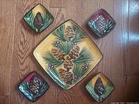 Top-down view of all five ceramic dishes arranged on wood floor, showing pinecone and pine needle designs on yellow and dark red backgrounds