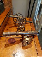 Two vintage brass replica field artillery cannons displayed on a wooden surface, showing overall form and style from side angle.
