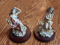 Two resin garden nymph sculptures on wooden bases on wooden floor.