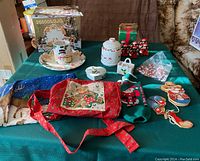 Full view of assorted Christmas holiday items arranged on a dark green tablecloth including box, porcelain trinket, ceramic plate and mug, fabric items, candy canes, and figurines.