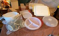 Wide photo of assorted vintage Tupperware items including white plastic containers, round divided tray, microwave splatter guards, cheese slicer and egg holders on tabletop