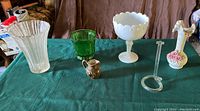 Full view of assorted vintage glassware on green tablecloth, including milk glass candy dish, green beaded glass planter, clear fluted vase, small decorated ceramic vase, creamer pitcher, and plastic stand.