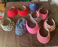 Nine visors and one hat laid out on a brown surface for clear viewing. Various colors and floral and checkered patterns represented.