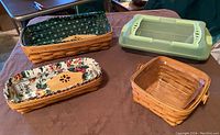 Three assorted woven Longaberger baskets with cloth liners and plastic protectors, plus one cupcake pan with lid on a table.