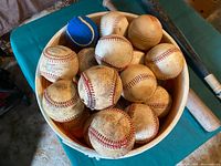 Bucket filled with approximately 20+ baseballs showing various degrees of wear, some with dirt marks and surface discoloration. A wooden baseball bat is lying beside the bucket with visible surface wear.