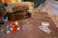 Photo showing two brown rectangular Pyrex glass casserole dishes, one with metal cradle, a clear Pyrex lid, assorted plastic pourers, a metal sifter, and other kitchen items.
