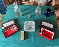 Full lot on green tablecloth showing all items: two glass decanters, their stoppers, jigger, strainer, ashtray, airplane bottle, blue cup, and small green box.