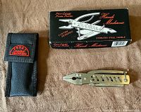 Photo of the multi-tool closed, next to the black sheath with red embroidery and original box.