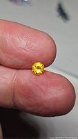 Close-up photo of a round brilliant cut golden yellow sapphire held between thumb and forefinger, showing size (approximately 5mm) and color.