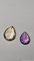 Two pear-shaped gemstones laid on grey background, showing deep purple amethyst on right and golden citrine on left, both faceted and polished.