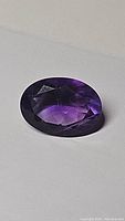 Top angle photo of deep purple oval cut amethyst on white background showing facets and internal characteristics.