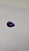 Close-up photo of a deep purple pear-shaped amethyst gemstone with checkerboard cut facets on white background.