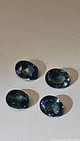 Four oval cut multicolored blue-green sapphires displayed on a flat surface showing dimensions and faceting details.
