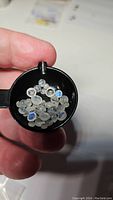 Close-up image showing a small black scoop containing approximately 34 small round cabochon rainbow moonstone gemstones with blue flash, held between fingers.