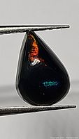 Close-up view of the pear cabochon black opal held with tweezers showing vibrant patches of red and blue play-of-color.