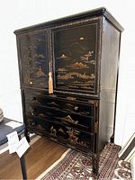 Front view of Asian lacquered dresser with two doors on top and three drawers below, decorated with gold painted scenes.