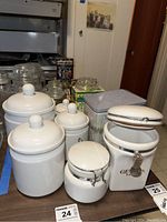 Four white ceramic canisters with lids, one with metal clasp and hinged lid that does not stay closed. Also visible are various glass canisters and metal tins.