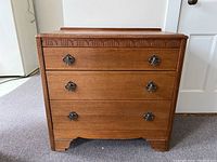 Front view of wooden Lebus chest of drawers showing three drawers with metal handles and carved top trim.