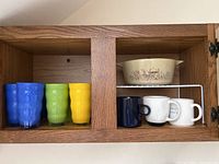 Wooden cabinet with three colored glasses (blue, green, yellow) and three coffee mugs with a Pyrex serving bowl on a rack above