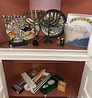 Shelf with three metal menorahs, colorful decorative plates, small metal figurines, and ceramic matzo plate with Hebrew lettering.