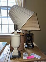 Two table lamps on a wooden surface near a window. One lamp has a cream pleated shade that is damaged. The other lamp has two separate brown lamp shades, one of which shows damage.