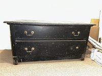 Front view of a low black wooden chest of drawers with two drawers, showing brass drop handles and scratches on the surface.