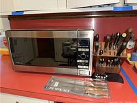 Panasonic microwave oven with digital panel next to wooden knife block holding knives and scissors, along with a sealed 3-piece knife set in original packaging.