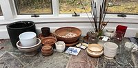 Wide view of all the terra cotta pots, ceramic pots with vintage frog designs, glass vases, shells, and small bowls arranged on a stone surface near a window.