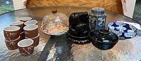 Photo showing seven brown saki cups with white bamboo leaf pattern, tea canister, black soup bowl, acrylic covered dish, blue and white bowl, and black elephant figurine on glass table.