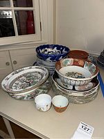 Shows collection of Asian style porcelain plates, bowls and small cups arranged on table; various patterns and colors including blue, orange, green, and white. Condition shows chips and repairs.