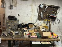 Wide view of basement workbench with assorted vintage bicycle repair tools, boxed hardware and parts, cables, manuals, and bicycle maintenance books scattered on the surface.