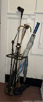 Metal umbrella stand with ornate top and circular base holding golf clubs, bamboo canes, and umbrellas including one with a blue handle.