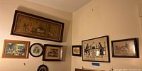 Photo showing multiple framed artworks hung on a wall including landscapes, a needlepoint silhouette, and a print with religious text.