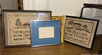 Group shot of four framed wall art pieces: Picasso 'Dove' print and three needlepoint samplers with inscriptions and house/tree designs.