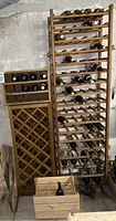 Photo showing the two wooden wine racks and the wooden wine crate, placed next to each other against a concrete wall. Several wine bottles are inserted in the racks for size reference; bottles are not included.