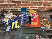 Wide view of lot showing dolls, Chinese checkers, checkers and dominoes boxes, wooden peg solitaire game on glass table with brick background.