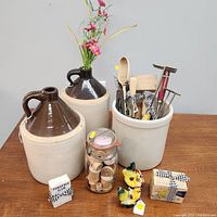 Photo showing two large antique stoneware crock jugs with dark brown tops and handles, a crock filled with antique kitchen utensils, a glass jar with wooden spools, two small ceramic bird figurines, and a small box tied with ribbon.