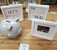 Photo showing a white ceramic piggy bank with pink polka dots and three white decorative frames, two with snuggle themed text and one with an ultrasound image, all arranged on a wooden table.
